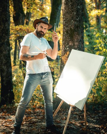 Téléchargez les photos : Artiste hipster dans un chapeau créant de l'art dans les bois. Concept d'art. Peinture dans la nature. Commencez une nouvelle image. Peintre avec chevalet et toile - en image libre de droit