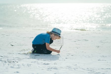 Téléchargez les photos : Joyeux enfant dessinant sur un sable dans la mer d'été. Un gamin qui s'amuse à la plage. Vacances d'été et enfants actifs. Voyage et concept d'aventure estivale. Enfant écrire dans le sable en vacances d'été - en image libre de droit