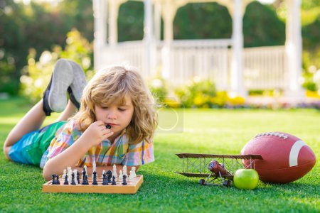 Téléchargez les photos : Jeux d'été et activités de plein air pour les enfants. Développement précoce. Garçon pense aux échecs dans le parc d'été. Le concept d'apprentissage et de croissance des enfants. Jeu en plein air, passe-temps des enfants et mode de vie - en image libre de droit