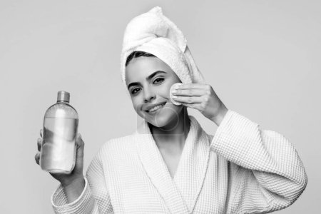Téléchargez les photos : Joyeux jeune femme en peignoir avec serviette sur le visage de nettoyage de la tête avec de l'eau micellaire et coton isolée sur rose. Fille avec de l'eau micellaire - en image libre de droit