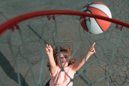 Foto de Lindo niño pequeño en uniforme de baloncesto saltando con pelota de baloncesto para el tiro. Niño feliz jugando al basket ball en el parque infantil - Imagen libre de derechos