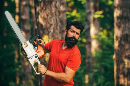 Foto de Un leñador profesional sosteniendo motosierra en el bosque. Un joven guapo con barba lleva un árbol. Cosecha de madera. Explotación - Imagen libre de derechos