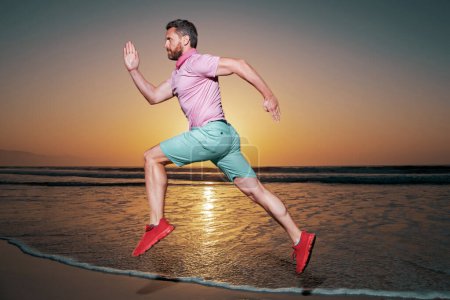 Foto de Atlético joven sano hombre corriendo al amanecer junto al mar. Modelo masculino en ropa deportiva ejercitando al aire libre sobre fondo de mar puesta de sol - Imagen libre de derechos