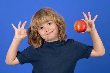 Photo for Portrait of a child with ok sign gesture hold a red apple on blue studio isolated background - Royalty Free Image