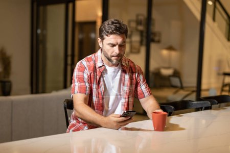 Photo for Handsome cheerful man indoors at home on sofa using mobile phone, drink coffe. Attractive caucasian man resting on sofa in apartment at night texting on call phone, surfing internet on smartphone - Royalty Free Image