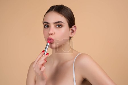 Foto de Gorgeous young woman in stylish makeup. Girl posing with new lipstick in studio. Sexy girl rouging her lips. Sexy model holding red lipstick. Applying lipstick cream balm to mouth. Red is trending - Imagen libre de derechos