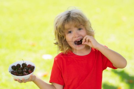 Photo for Happy little child with cherry outdoors. Kid picking and eating ripe cherries in summer park. Child holding fresh fruits. Healthy organic berry cherry fruit. Summer background with green grass - Royalty Free Image