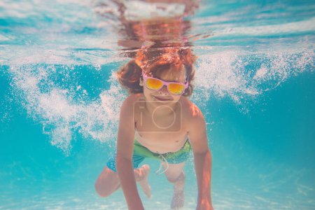 Téléchargez les photos : Le gamin nage sous l'eau dans la piscine d'été. Concept vacances d'été pour enfants. Les enfants drôles font face sous l'eau. Des éclaboussures d'enfants dans la piscine. Sports nautiques d'été. Vacances d'été avec enfant - en image libre de droit