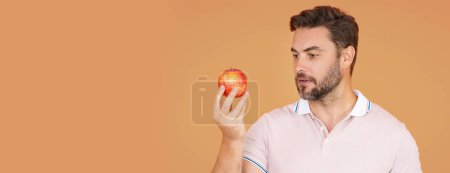 Téléchargez les photos : Homme d'âge moyen piquant pomme, mode de vie sain. Homme tient un portrait frais de studio de pomme sur fond isolé beige. Une nourriture saine. Fruits pour des dents saines. Un homme tenant une pomme. Bannière, espace de copie - en image libre de droit