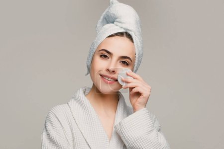 Foto de Mujer en toalla de spa con crema facial y almohadilla de algodón aislada sobre fondo gris - Imagen libre de derechos