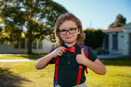 Téléchargez les photos : Heureux écolier souriant dans des lunettes avec le pouce levé va à l'école pour la première fois. Enfant avec cartable. Enfant à l'extérieur de l'école - en image libre de droit