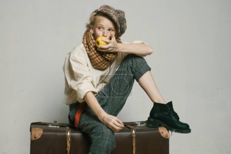 Photo for Travel with old suitcase. Homelss. old fashioned child in beret eat apple. retro fashion model. Vintage suspender. teen girl in retro male suit. street kid with dirty face. vintage english style. - Royalty Free Image