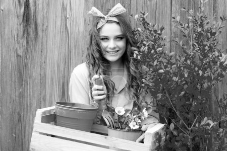 Foto de Plantando flores. Chica bastante sonriente sosteniendo herramientas de jardín en el fondo de madera. Chica en una chaqueta amarilla sosteniendo maceta en el jardín - Imagen libre de derechos