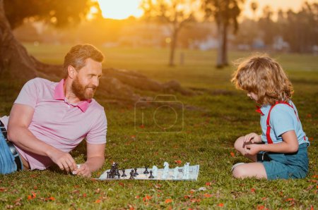 Téléchargez les photos : Fils étendu sur l'herbe et jouant aux échecs avec son père. Le petit garçon joue aux échecs avec ses parents. Développement cognitif. Jeu intellectuel pour enfants. Enfants intelligents - en image libre de droit