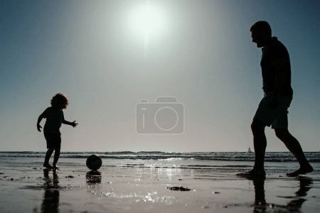 Téléchargez les photos : Père et fils jouent au football ou au football sur la plage. Papa avec enfant garçon un jour d'été - en image libre de droit