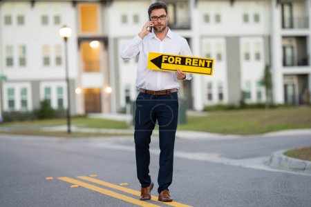 Photo for Successful real estate agent in a suit holding for rent sign near new apartment. Real estate agent with home loan contract, renting home. Realtor or real estate agent shows board for rent - Royalty Free Image