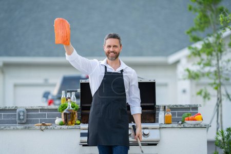 Photo for Grill cook. Chef in cook apron with BBQ cooking tools. Barbecue and grill. Chief cook hold salmon fillet with utensils for barbecue grill. Barbeque on holiday picnic. Man grilling fish on BBQ - Royalty Free Image