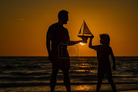 Téléchargez les photos : Père et fils jouant avec le bateau jouet sur la mer. Silhouette coucher de soleil de père et fils rêvant en croisière. Rêver de voyager. Père et fils se sentent aventureux en croisière. Concept de voyage et d'aventure - en image libre de droit