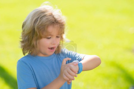 Téléchargez les photos : L'enfant vérifie la montre intelligente, regarde le temps et se dépêche. Portrait d'enfant vérifiant l'heure dans le parc de printemps à l'extérieur. Gros plan visage enfant jouant à l'extérieur dans un parc d'été - en image libre de droit