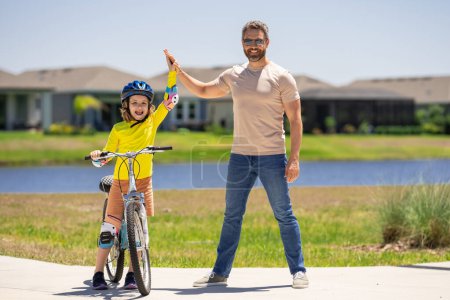 Photo for Father support son. Happy Fathers day. Father and son on the bicycle. Father and son riding a bike in summer park. Kid learning to riding bicycle. Father and son riding bikes wearing helmets - Royalty Free Image