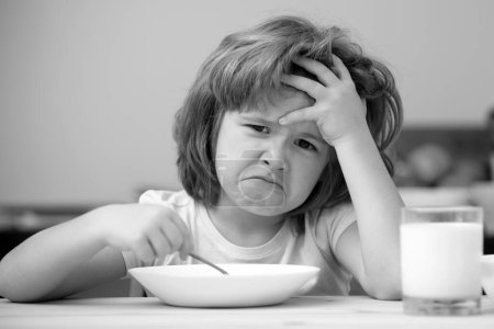 Foto de Niño desconcentrado almorzando sopa. Infeliz niño caucásico sentarse a la mesa en la cocina casera no tienen apetito. Niño molesto se niega a comer cereales orgánicos con leche - Imagen libre de derechos