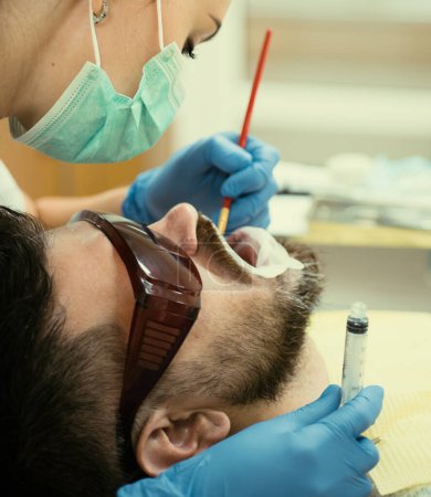 Foto de Examine al hombre joven por dentista sobre fondo borroso claro. Dentista femenina revisando dientes de paciente con espejo en clínica dental moderna. Hombre barbudo con dientes examinados en dentistas - Imagen libre de derechos