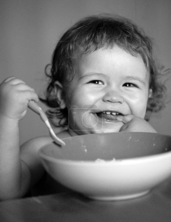 Foto de Bebé sonriente comiendo comida. Divertido bebé comiendo comida él mismo con una cuchara en la cocina. Nutrición saludable para los niños. Lanzamiento niño comer alimentos - Imagen libre de derechos