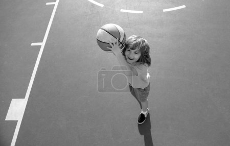 Photo for Cute smiling boy plays basketball, outdoor on playground. Child shooting Basketball ball and playing basketball, lower view wide angle - Royalty Free Image