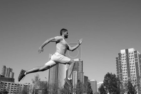 Téléchargez les photos : Toute la longueur de l'homme en bonne santé courir et le sprint à l'extérieur. Modèle masculin en vêtements de sport faisant de l'exercice en plein air dans le parc de la ville. Concept de sport urbain - en image libre de droit