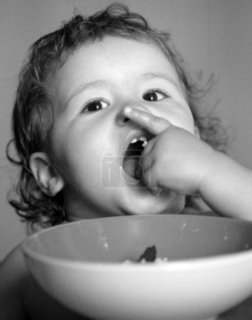 Téléchargez les photos : Petit bébé drôle dans la cuisine manger avec les doigts de la plaque. Enfant drôle visage gros plan - en image libre de droit