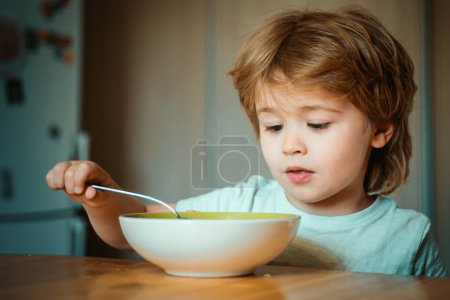 Foto de Sonriendo feliz adorable bebé comiendo puré de frutas en la cocina. El niño en la cocina en la mesa comiendo. Alegre bebé niño come comida sí mismo con cuchara - Imagen libre de derechos