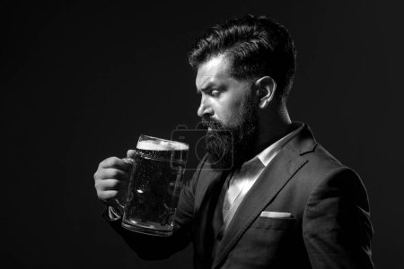 Téléchargez les photos : Un homme en costume classique buvant de la bière. Barbu gars en tenue de travail semble heureux et satisfait. Portrait profil de l'homme avec haut verre de bière levé sur fond noir - en image libre de droit