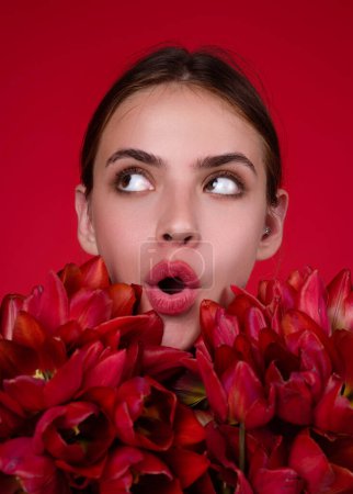 Téléchargez les photos : Surprised woman face with flowers. Beautiful sensual woman hold tulips bouquet, studio portrait on red background. Close up beauty woman face near tulips bouquet - en image libre de droit