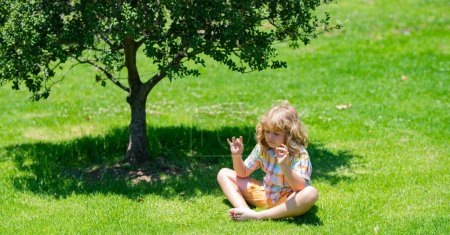 Téléchargez les photos : Enfant méditant en position lotus sur l'herbe, relaxant sur la nature estivale. Petit enfant heureux médite sur l'herbe verte le jour de l'été. Concept de liberté - en image libre de droit