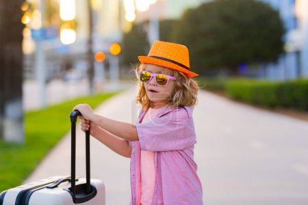 Foto de Divertido viajero infantil. Niño viajero sosteniendo maleta al aire libre. Turista chico teniendo alegre viaje de vacaciones - Imagen libre de derechos