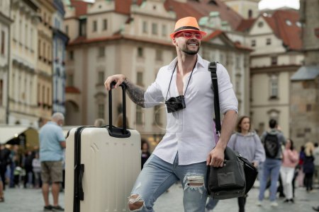 Foto de Hombre de turismo en jeans sombrero de moda y gafas de sol con maleta caminando por la calle. Hombre con bolsa de viaje listo para viajar en sus vacaciones - Imagen libre de derechos