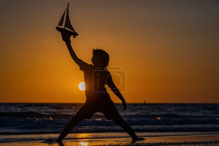 Photo for Silhouette of kid playing with toy seailing boat on sunset sea. Child playing with toy boat in sea. Happy holiday by the sea. Little sailor. Kid dreaming about sailing - Royalty Free Image