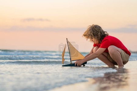 Téléchargez les photos : Petit garçon jouant avec un bateau jouet dans l'eau de mer. Joyeuses vacances au bord de la mer. Enfant rêvant de naviguer. Enfant heureux jouant avec un voilier jouet. Vacances d'été. Mignon petit enfant tenir jouet voilier - en image libre de droit