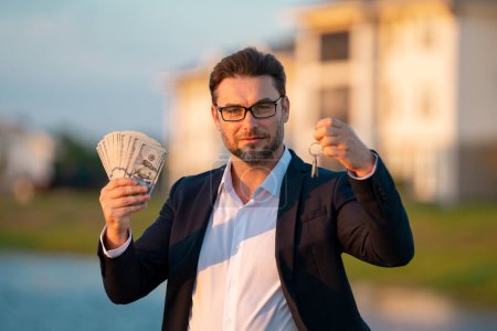 Photo for Handsome real estate agent holding money dollars and house keys. Man real estate agent in business suit presenting the house for sale. New apartment after buying real estate - Royalty Free Image