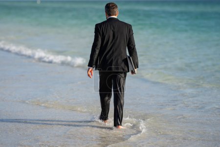 Foto de Vista trasera del hombre de negocios espalda en traje en agua de mar en la playa. Descanso de verano, concepto de gente de negocios. Hombre de negocios remoto trabajando en línea. Sueño para los trabajadores de oficina de vacaciones de verano - Imagen libre de derechos