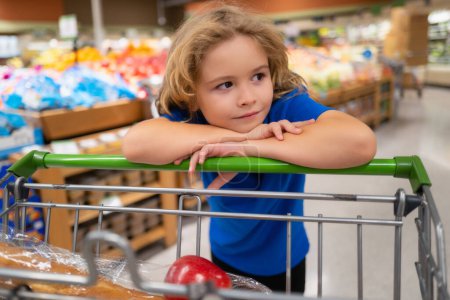 Téléchargez les photos : Enfant avec caddie à l'épicerie. Enfant achetant des fruits au supermarché. Le petit garçon achète des légumes frais à l'épicerie. Un gamin qui choisit des légumes. Aliments sains - en image libre de droit