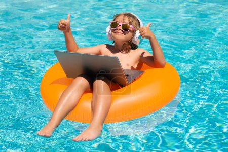 Téléchargez les photos : Enfant se détendre dans la piscine, en utilisant un ordinateur portable dans l'eau d'été. Enfant étude en ligne ou de travailler sur la plage de la mer tropicale. Concept de technologie pour la vie. Voyage d'été et concept d'affaires - en image libre de droit