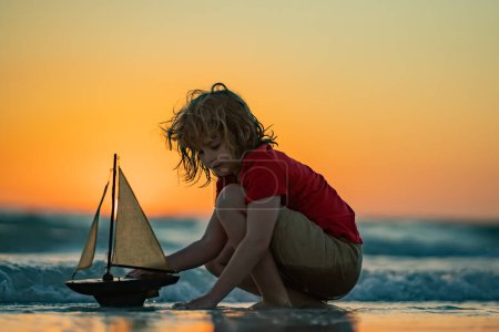Téléchargez les photos : Petit garçon blond mis bateau jouet dans les vagues de la mer à la plage pendant les vacances d'été. Vacances d'enfance et d'été en famille. Rêve en voyage. Visite guidée en voilier. Concept de rêve enfants - en image libre de droit