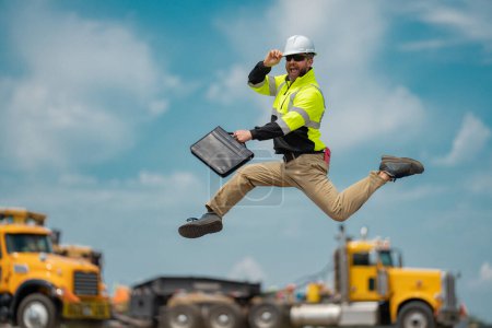 Photo for Hispanic builder excited jump and run on site construction. Excited builder construction worker in a safety helmet jumping in front of the trucks. Excited crazy builder man in helmet jump outdoor - Royalty Free Image