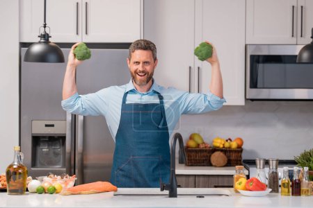 Photo for Man in chef apron with broccoli on kitchen. Man cooking vegan healthy salad in kitchen. Millennial man at modern kitchen with vegetables, prepare fresh vegetable - Royalty Free Image