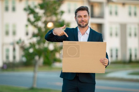 Photo for Man in business suit holds the sign, blank card. Placard ready for your product. Sign to your text. Business man showing blank sign board outdoor. Empty blank board. Area for advertising - Royalty Free Image