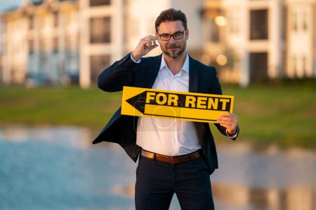 Photo for Successful real estate agent in a suit holding for rent sign near new apartment. Real estate agent with home loan contract, renting home. Realtor or real estate agent shows board for rent - Royalty Free Image
