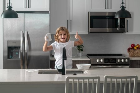 Foto de Divertidos chicos gemelos ayudando en la cocina con lavar los platos. Niños divirtiéndose con tareas domésticas. Niño lavando platos en el interior de la cocina. Niño ayudando a sus padres con las tareas domésticas - Imagen libre de derechos