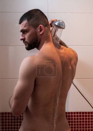 Foto de Cuidado de la piel y del cuerpo. Joven musculoso duchándose lavándose el cuerpo, espalda y hombros. Un tipo guapo parado en el baño bajo gotas de agua caliente. Rutina de limpieza higiénica en el baño - Imagen libre de derechos