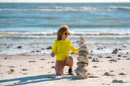 Téléchargez les photos : Méditation estivale pour enfants. Enfant avec des pierres zen sur la plage de la mer, méditation, spa et harmonie. Calme concept d'équilibre. Les enfants jouent sur la plage. Calmant enfant et tour de pierre contre la mer - en image libre de droit